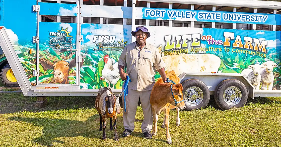 Life on the Farm (LOTF) coordinator, Chris Sanders, standing with a cow and a goat in front of the LOTF trailer.