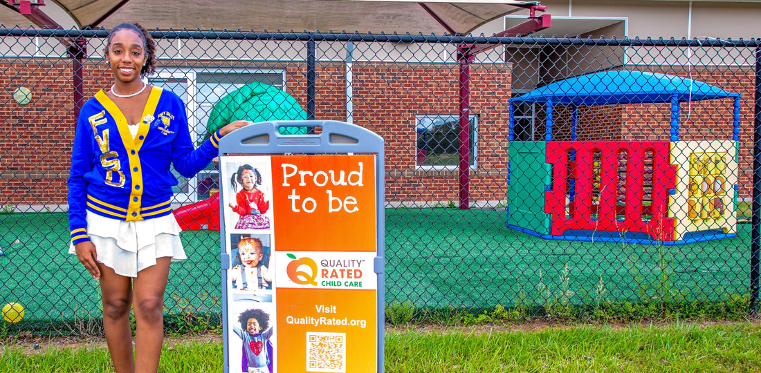 FCS student standing next to a Qality Rated Childcare sign in front of the Family Development Center's playground